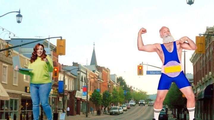 A red-haired woman pointing at her laptop and a bearded senior flexing in a blue wrestling singlet, with Milton in the background